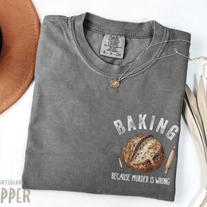 Op de afbeelding: Een grijs Comfort Colors T-shirt met de tekst "BAKING BECAUSE MURDER IS WRONG" en een afbeelding van een brood, garde en deegroller. Een bruine hoed en ketting zijn ook zichtbaar.