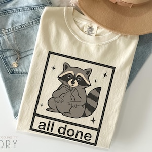 May include: Ivory-colored t-shirt with a raccoon graphic inside a black square, with the words "all done" below. The shirt is folded on top of blue jeans and a tan hat.