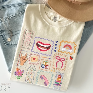 Puede incluir: Camiseta de color marfil con gráficos coloridos con temática de terapia del habla. El diseño incluye una boca, un arcoíris, un signo de mano y otras ilustraciones. La camiseta está colocada sobre unos vaqueros azules y un sombrero marrón.