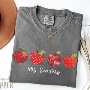 Op de afbeelding: Een grijs Comfort Colors T-shirt met vier geappliqueerde rode appels. De appels hebben verschillende patronen en texturen. De tekst "Mrs. Sanders" staat eronder gedrukt. Een zilveren ketting met een hanger ligt op het shirt.