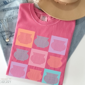 May include: A pink Comfort Colors t-shirt with a grid pattern of colorful pots. The shirt is folded on top of a pair of blue jeans and a tan hat. The text "Comfort Colors 1717 Crunchberry" is printed on the bottom left.