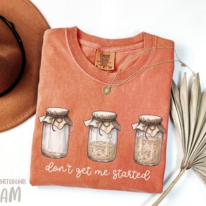 May include: Orange Comfort Colors t-shirt with a graphic of three jars, each with a different stage of fermentation. The text "don't get me started" is below the jars. A gold necklace and a brown hat are also in the image.
