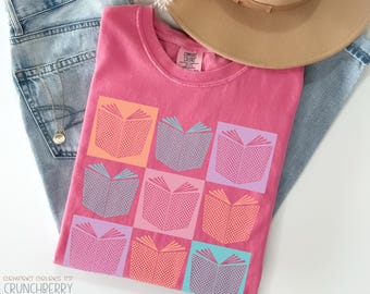 May include: A pink Comfort Colors t-shirt with a grid pattern of open books in various colors. The shirt is folded on top of a pair of blue jeans and a tan hat. The text "Comfort Colors 1717 Crunchberry" is visible.