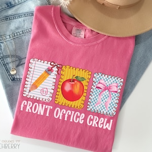 May include: A pink Comfort Colors t-shirt with the text "FRONT OFFICE CREW" and three graphic designs: a pencil, a red apple, and a pink bow. The shirt is laid on top of blue jeans and a tan hat.