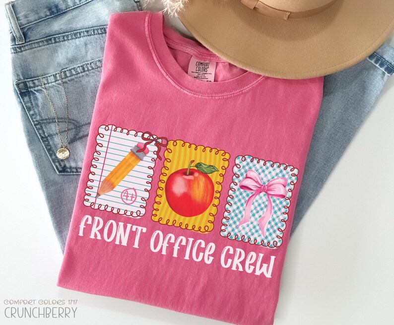 May include: A pink Comfort Colors t-shirt with the text "FRONT OFFICE CREW" and three graphic designs: a pencil, a red apple, and a pink bow. The shirt is laid on top of blue jeans and a tan hat.