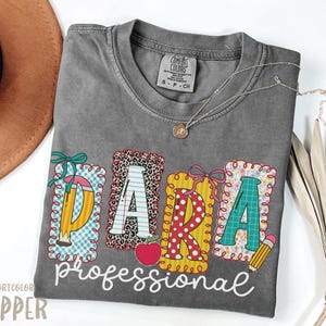 May include: A gray Comfort Colors t-shirt with the word "PARA" in colorful, decorative letters, and the word "professional" in white script. The letters are designed with patterns like leopard print, polka dots, and floral prints. A pencil and apple are also featured.