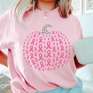 May include: Pink sweatshirt featuring a pumpkin design made of pink ribbons, with a green stem. The sweatshirt is a light pink color. The design is a symbol of breast cancer awareness.