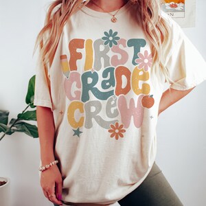 May include: A cream colored t-shirt with the text "First Grade Crew" in colorful, retro-style lettering. The shirt features a pencil, flowers, and stars. The model is wearing a size L.