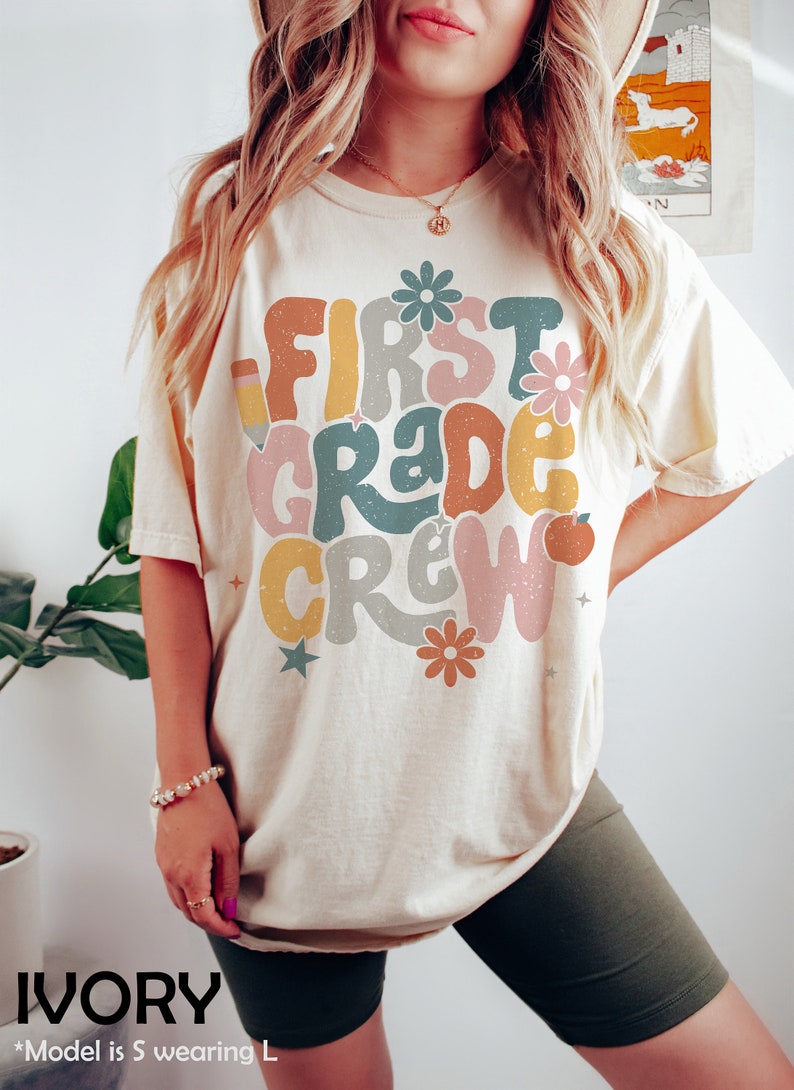May include: A cream colored t-shirt with the text "First Grade Crew" in colorful, retro-style lettering. The shirt features a pencil, flowers, and stars. The model is wearing a size L.