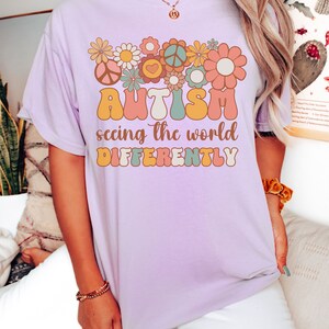May include: A lavender t-shirt with a graphic design of colorful flowers and peace signs. The text "AUTISM seeing the world DIFFERENTLY" is printed on the shirt.