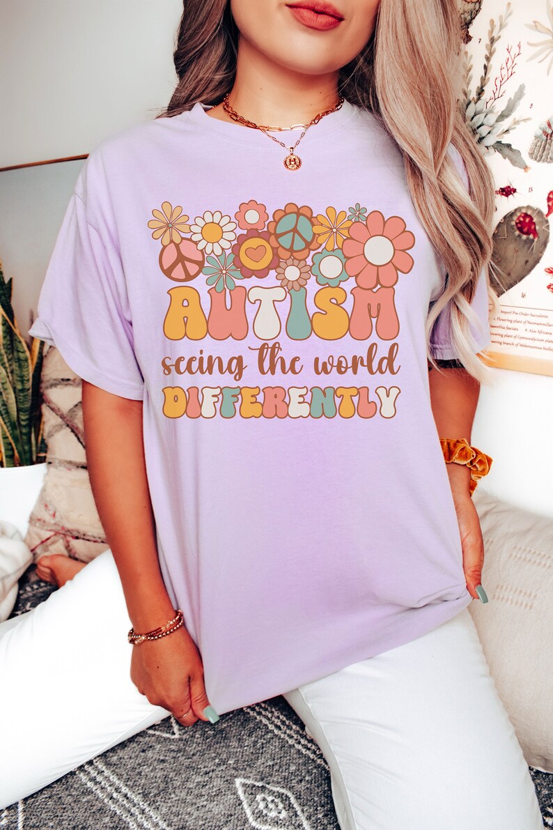 May include: A lavender t-shirt with a graphic design of colorful flowers and peace signs. The text "AUTISM seeing the world DIFFERENTLY" is printed on the shirt.
