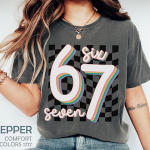 May include: Dark gray t-shirt with the numbers "67" in a rainbow gradient design, the word "six" above, and "seven" below. The background is a black and white checkered pattern. The shirt has the word "Pepper" and "Comfort Colors 1717" printed on it.