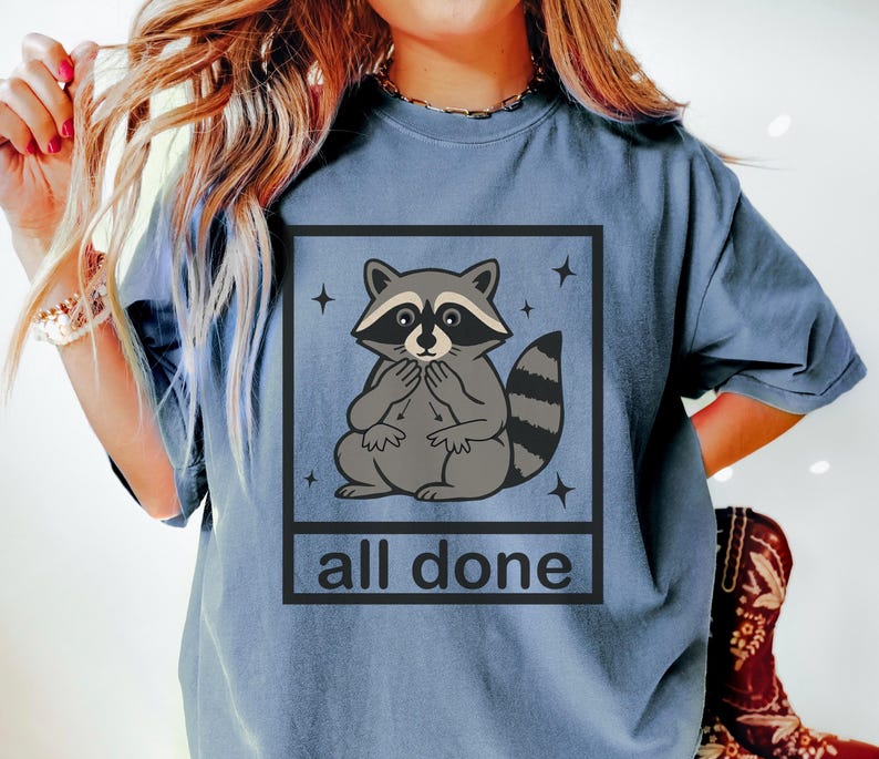 May include: A blue t-shirt featuring a cartoon raccoon inside a black square with the text "all done". The raccoon is gray and white with a black mask and striped tail. Four small stars surround the square.