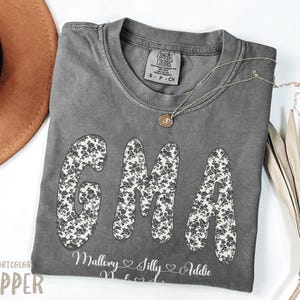 May include: A grey Comfort Colors t-shirt featuring the letters "GMA" in a floral design. The names Mallory, Silly, and Addie are printed below. A gold necklace and a brown hat are also visible.
