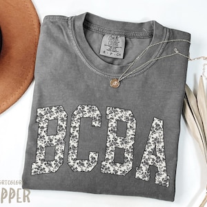 Op de afbeelding: Een grijs Comfort Colors T-shirt met de letters "BCBA" in een bloemenpatroon. Het shirt heeft een ronde hals en korte mouwen. Een gouden ketting met een hanger is over het shirt gedrapeerd. Een bruine hoed is zichtbaar op de achtergrond.