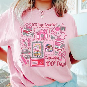 May include: Pink sweatshirt with a colorful graphic celebrating the 100th day of school. The design includes books, a school bus, a rainbow, and the text "100 Days Smarter!" and "Happy 100th Day!"