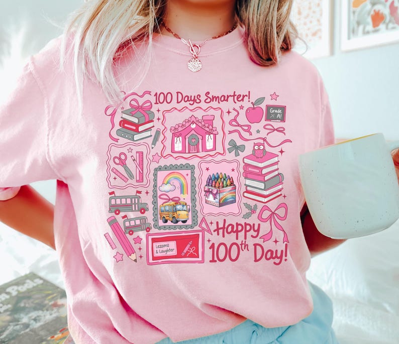 May include: Pink sweatshirt with a colorful graphic celebrating the 100th day of school. The design includes books, a school bus, a rainbow, and the text "100 Days Smarter!" and "Happy 100th Day!"