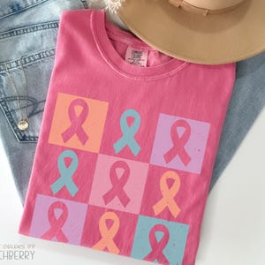 Puede incluir: Una camiseta rosa con un patrón de lazos coloridos en cuadrados naranjas, azules y rosas. La camiseta está colocada sobre unos vaqueros azules y un sombrero beige.