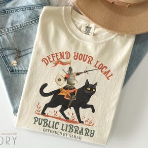 May include: Cream-coloured t-shirt featuring the text "Defend Your Local Public Library" and a graphic of a knight on a black cat. The knight is armed with a sword and shield. The shirt is shown with blue jeans and a beige hat.