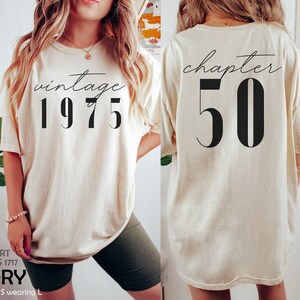 May include: Ivory oversized t-shirt with short sleeves. The front of the shirt has the text "vintage 1975" printed in black. The back of the shirt has the text "chapter 50" printed in black.