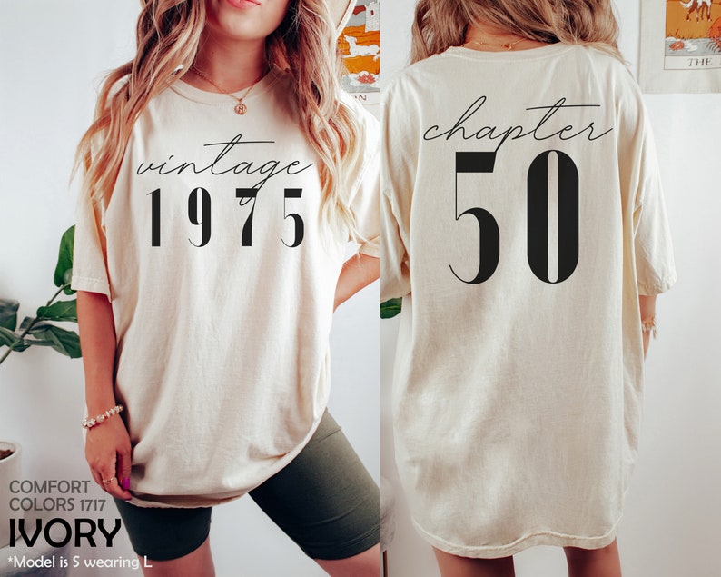 May include: Ivory oversized t-shirt with short sleeves. The front of the shirt has the text "vintage 1975" printed in black. The back of the shirt has the text "chapter 50" printed in black.