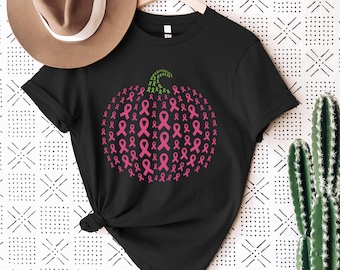 Op de afbeelding: Zwart T-shirt met een pompoenontwerp gemaakt van roze bewustzijns linten. De pompoensteel is groen. Het shirt hangt aan een houten hanger. Een bruine hoed is zichtbaar op de achtergrond.