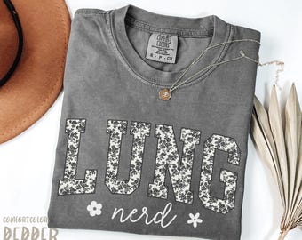 May include: A gray Comfort Colors t-shirt with the word "LUNG" in large, floral-patterned letters, and the word "nerd" in script below. A gold necklace and a brown hat are also visible.