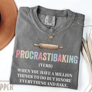 Op de afbeelding: Een grijs Comfort Colors T-shirt met de tekst "PROCRASTIBAKING" in pastelkleuren en een deegroller-afbeelding. Het shirt bevat ook de definitie van het woord. Een gouden ketting en een bruine hoed staan ook op de foto.