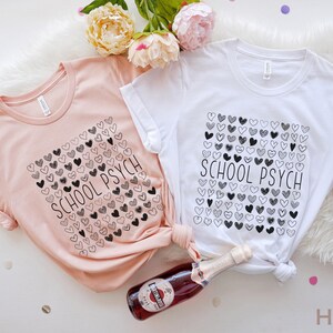 Pode incluir: Duas camisetas, uma p&ecirc;ssego e outra branca, com o texto "SCHOOL PSYCH" e um desenho de cora&ccedil;&atilde;o. Uma garrafa de vinho espumante e flores tamb&eacute;m est&atilde;o na imagem. As camisas est&atilde;o em uma superf&iacute;cie branca.