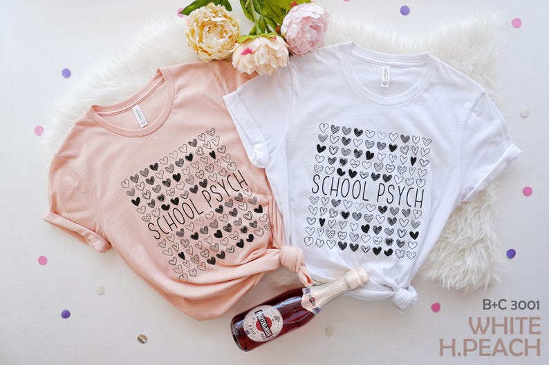 Pode incluir: Duas camisetas, uma p&ecirc;ssego e outra branca, com o texto "SCHOOL PSYCH" e um desenho de cora&ccedil;&atilde;o. Uma garrafa de vinho espumante e flores tamb&eacute;m est&atilde;o na imagem. As camisas est&atilde;o em uma superf&iacute;cie branca.