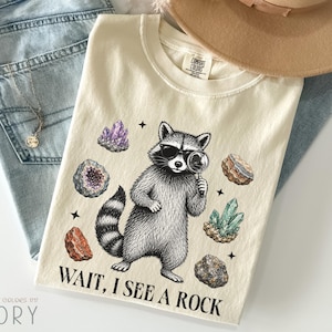 以下が含まれることがあります： サングラスをかけ虫眼鏡を持ったアライグマのグラフィックが描かれたアイボリーのTシャツ。周囲には様々な色のクリスタルや岩が配置されています。アライグマの下には「WAIT, I SEE A ROCK.」という文字がプリントされており、クルーネックのデザインです。