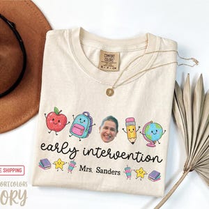 May include: An ivory t-shirt featuring a graphic print with a smiling apple, backpack, pencil, and globe, along with a photograph of a person's face. The text reads early intervention and Mrs. Sanders, accompanied by small illustrations of books, stars, and cups.