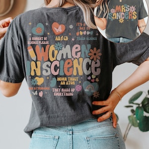 May include: A dark gray t-shirt with a colorful graphic that says "Women in Science" and includes a butterfly, hearts, stars, and rainbows. The text "A moment of science please", "It's a day to teach science", "ABCD", "Never trust an atom", and "They make up everything" are also included in the graphic. The t-shirt is being worn by a person with long brown hair.