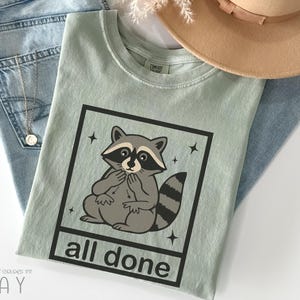 May include: A light green t-shirt featuring a cartoon raccoon inside a black square with the words "all done" below. The raccoon is gray and white with a black mask and striped tail. The shirt is laid on top of blue jeans and a tan hat.