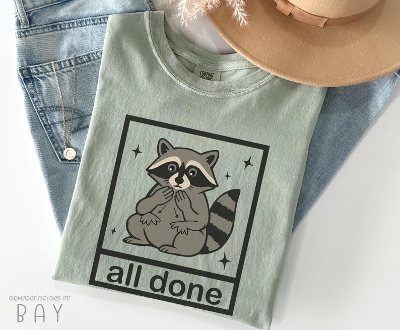 May include: A light green t-shirt featuring a cartoon raccoon inside a black square with the words "all done" below. The raccoon is gray and white with a black mask and striped tail. The shirt is laid on top of blue jeans and a tan hat.