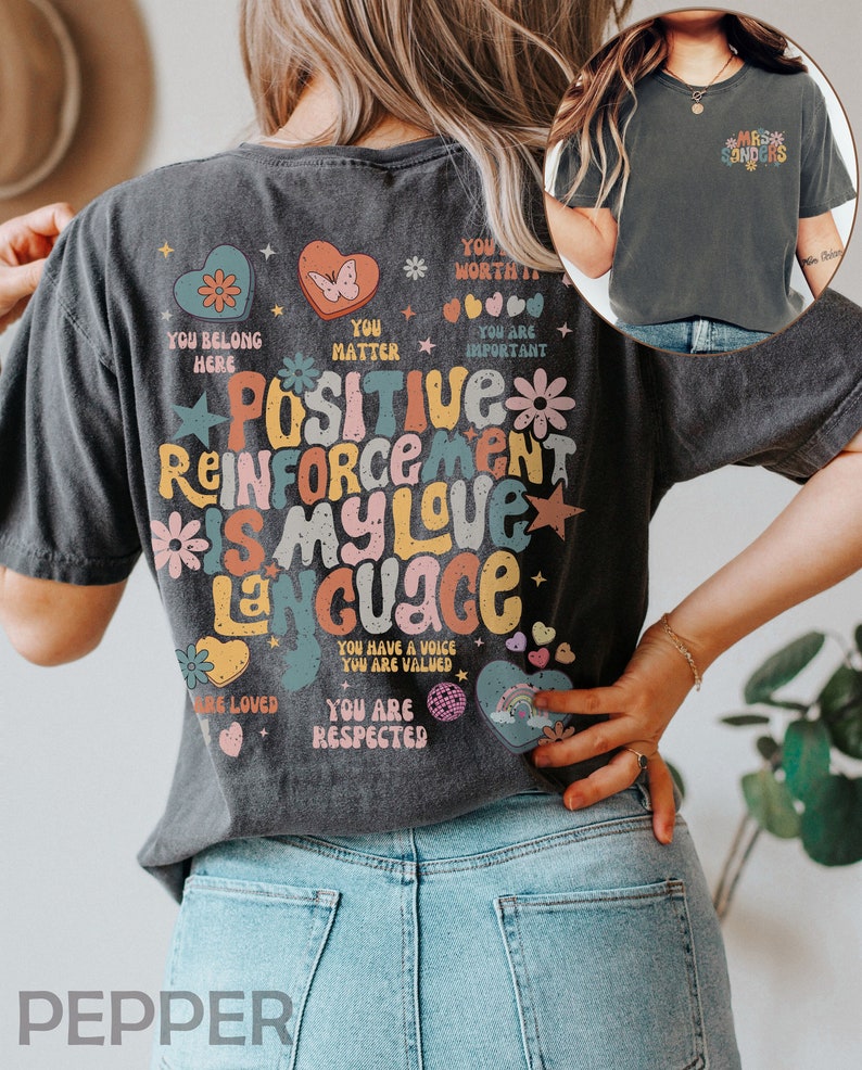 Pode incluir: Uma camiseta cinza escura com um design gr&aacute;fico colorido que diz "Positive Reinforcement is my love language". O gr&aacute;fico inclui cora&ccedil;&otilde;es, estrelas, flores e arco-&iacute;ris. O texto na camiseta tamb&eacute;m inclui frases como "You belong here", "You matter", "You are important", "You are loved", "You are respected", e "You have a voice, you are valued".