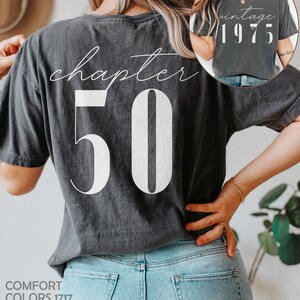 May include: A dark gray t-shirt with the text "chapter 50" printed on the back in white. The front of the shirt has the text "vintage 1975" printed in white.