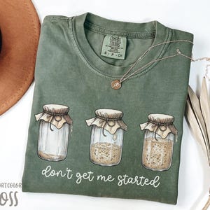 May include: Moss green Comfort Colors t-shirt featuring a graphic of three jars with paper lids, filled with a light brown substance. The text "don't get me started" is printed below the jars. A gold necklace is draped across the shirt.