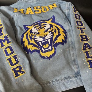 May include: A light blue denim jacket featuring a large glittery tiger head graphic in yellow, blue, and white on the back. The text MASON is printed above the tiger, with FOOTBALL and SEYMOUR on the sleeves. Clear rhinestones are scattered throughout.