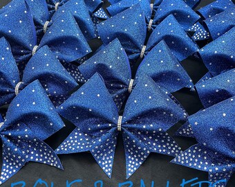 May include: A group of blue and silver cheer bows with rhinestones. The bows are arranged in a pile and have a sparkly finish. The text "BOWS & BALLERS" is visible at the bottom of the image.