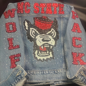 May include: A light blue denim jacket featuring red glitter lettering that reads NC STATE WOLF PACK. The back displays a large graphic of a wolf wearing a red hat, accented with scattered clear rhinestones across the entire garment surface.