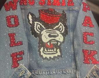 May include: A light blue denim jacket featuring red glitter lettering that reads NC STATE WOLF PACK. The back displays a large graphic of a wolf wearing a red hat, accented with scattered clear rhinestones across the entire garment surface.