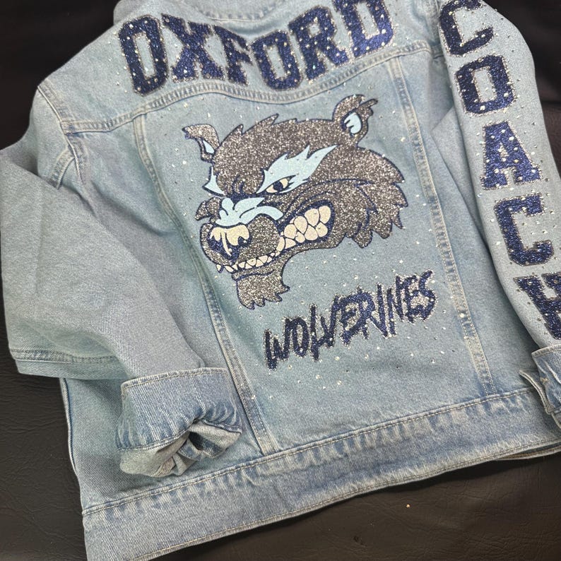 May include: A light blue denim jacket featuring a large glittery dark blue and silver wolverine mascot graphic on the back. The text OXFORD is at the top, WOLVERINES is below the mascot, and COACH is on the sleeve, all accented with clear rhinestones.