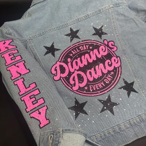 May include: A light blue denim jacket featuring a large circular graphic on the back with the pink text Dianne's Dance and the words All Day Every Day. The design includes black glitter stars and clear rhinestones. The sleeve features the name KENLEY.