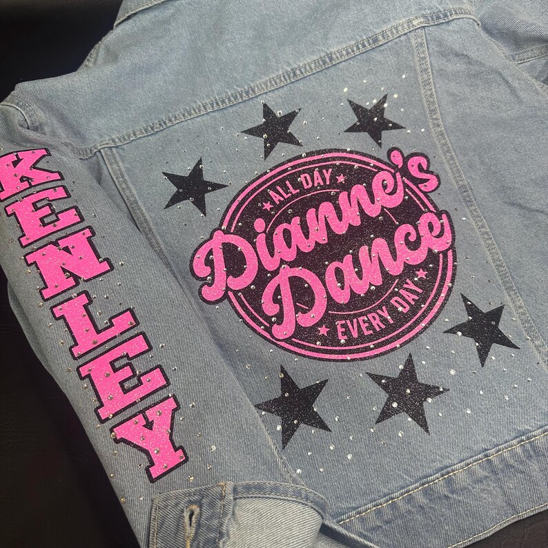 May include: A light blue denim jacket featuring a large circular graphic on the back with the pink text Dianne's Dance and the words All Day Every Day. The design includes black glitter stars and clear rhinestones. The sleeve features the name KENLEY.