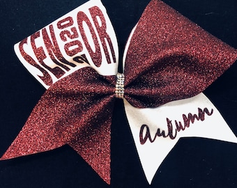 May include: A maroon glitter cheer bow with white accents. The bow features the word "SENIOR 2020" and the name "Autumn". A rhinestone band is in the centre of the bow.