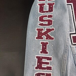 May include: A light blue denim jacket sleeve featuring the word HUSKIES written vertically in large, maroon glitter block letters with a white outline. The fabric is embellished with numerous small, clear rhinestones scattered around the text.