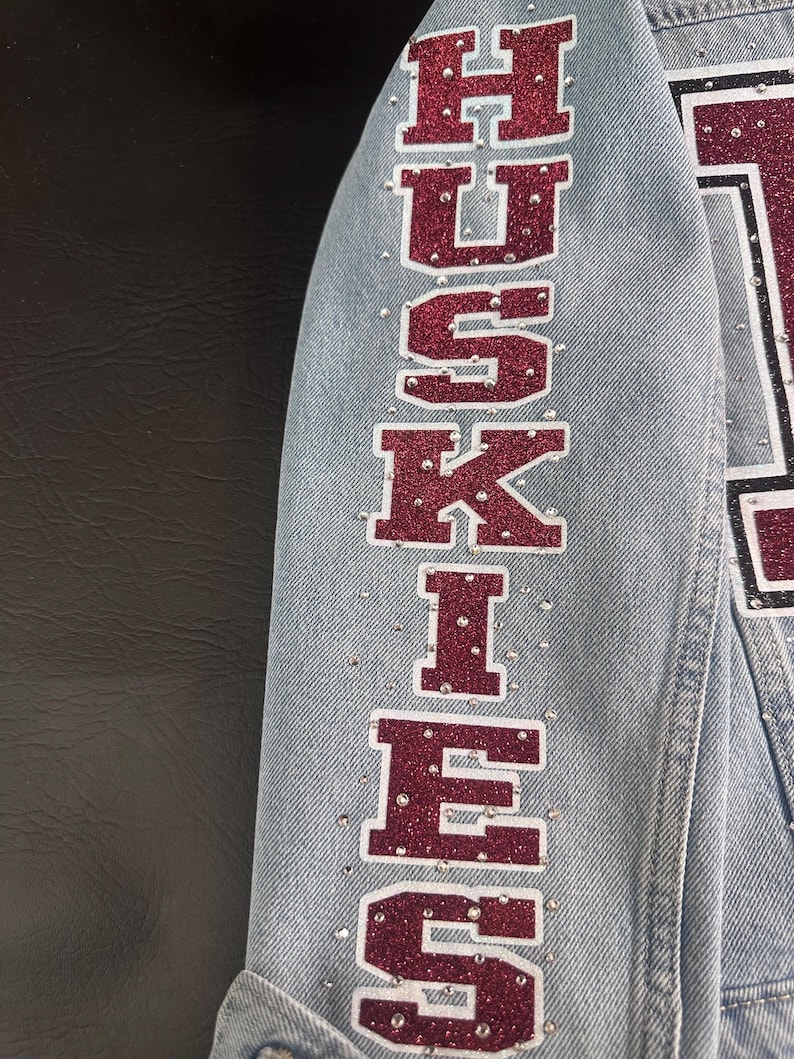 May include: A light blue denim jacket sleeve featuring the word HUSKIES written vertically in large, maroon glitter block letters with a white outline. The fabric is embellished with numerous small, clear rhinestones scattered around the text.