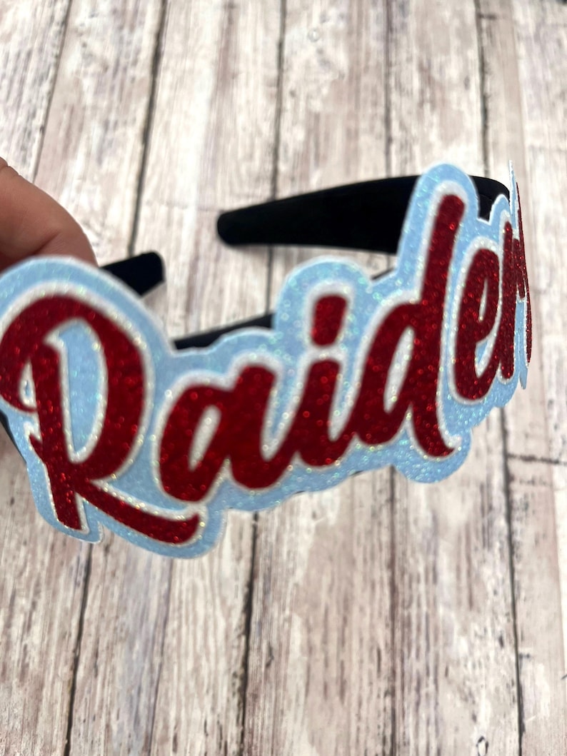 May include: A black headband with a red glitter "Raiders" word design on a light blue background.