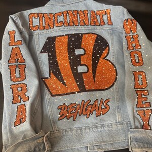 May include: A light blue denim jacket featuring orange glitter lettering that reads CINCINNATI, LAURA, WHO DEY, and BENGALS. A large orange glitter B logo is centred on the back, and the entire design is embellished with clear rhinestones.